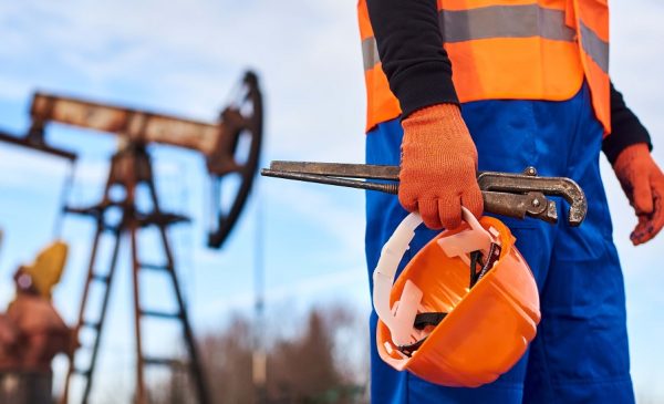 Cropped close-up snapshot of a man wearing blue overalls orange vest and gloves, holding a pipe wrench and a helmet on foreground, oil pump jack on background. Concept of petroleum industry.